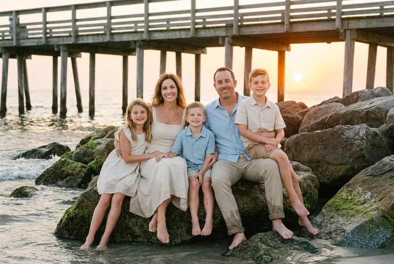 Family of five sitting on beach rocks under a pier at sunset