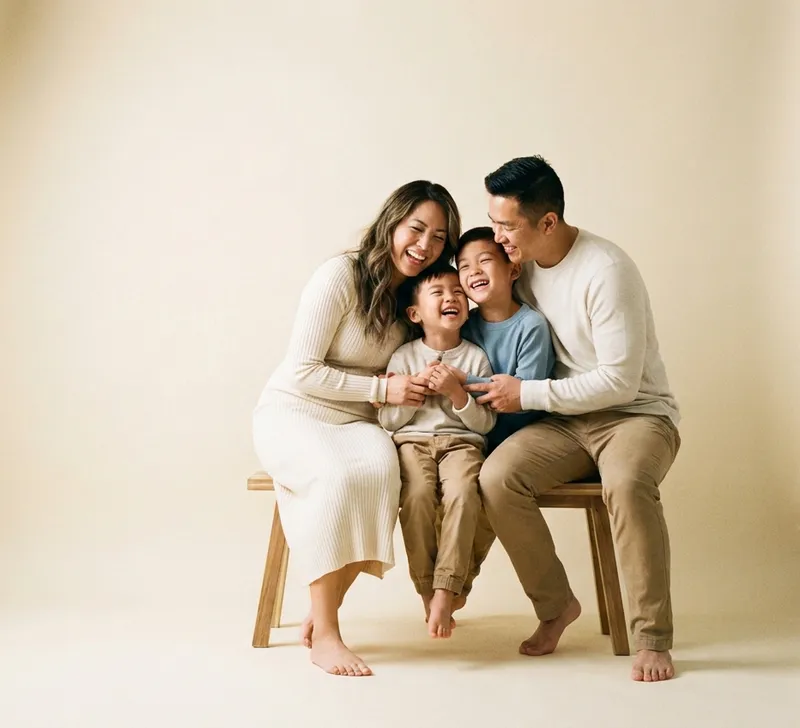 Family of four laughing together on a wooden bench against a cream studio backdrop