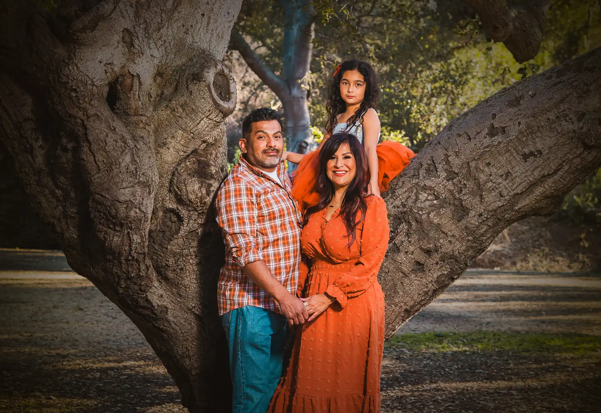 Family of three standing together under a large oak tree, mother wearing an orange dress