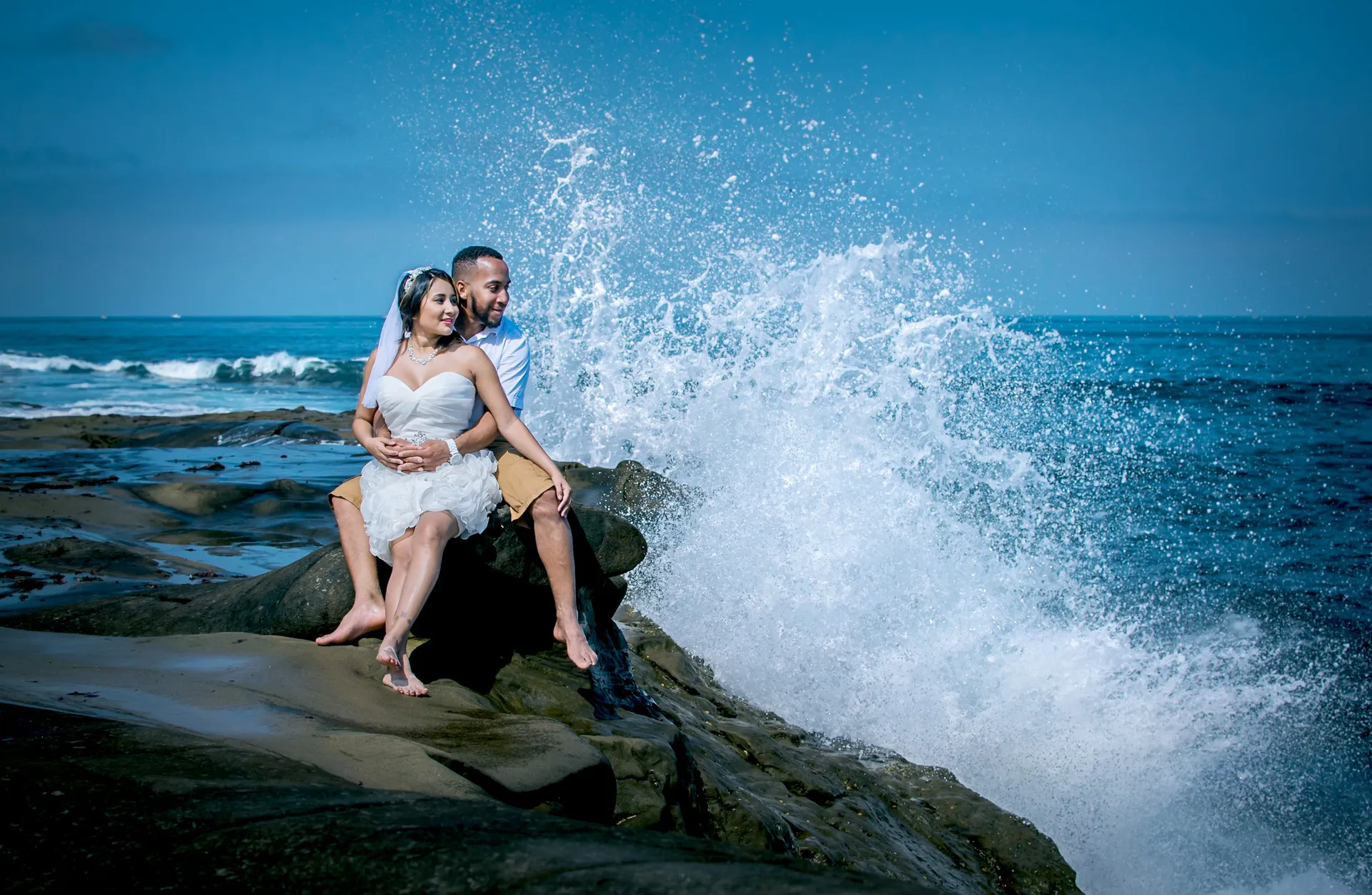 Bride and groom embracing on coastal rocks as a wave crashes behind them, Northridge wedding photography