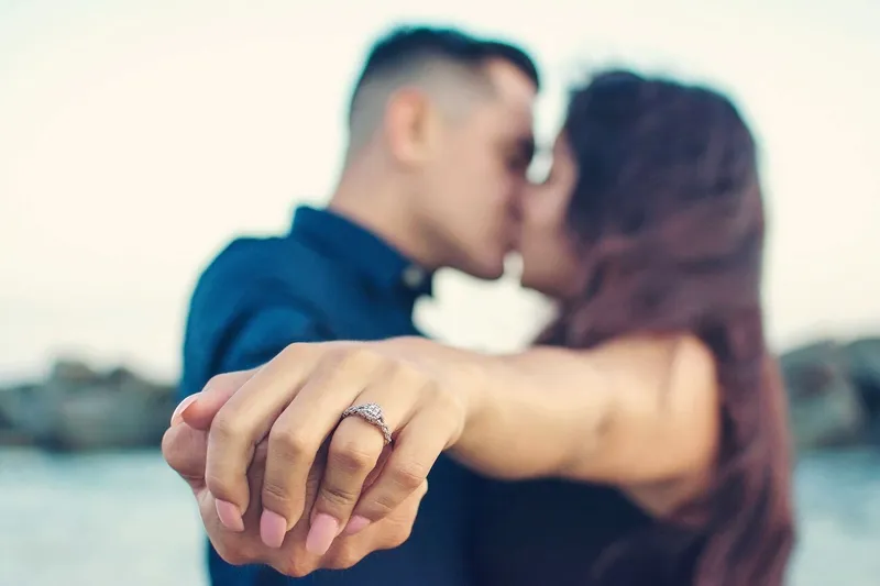 Close-up of an engagement ring with the couple kissing softly blurred in the background