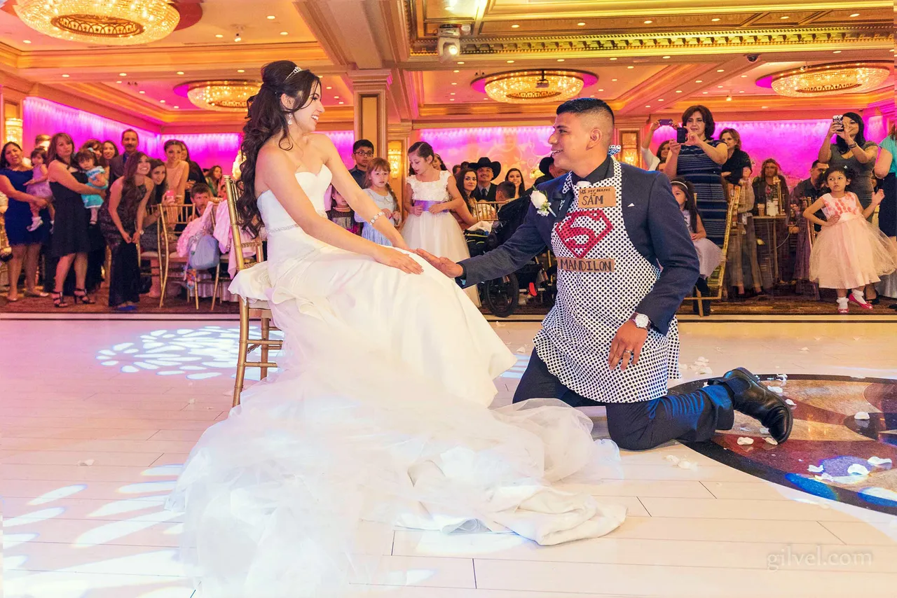 One person seated while another kneels wearing a novelty apron, reception guests nearby